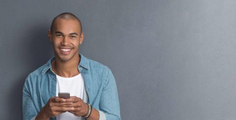 Smiling man in denim shirt holding a smartphone against a gray background.